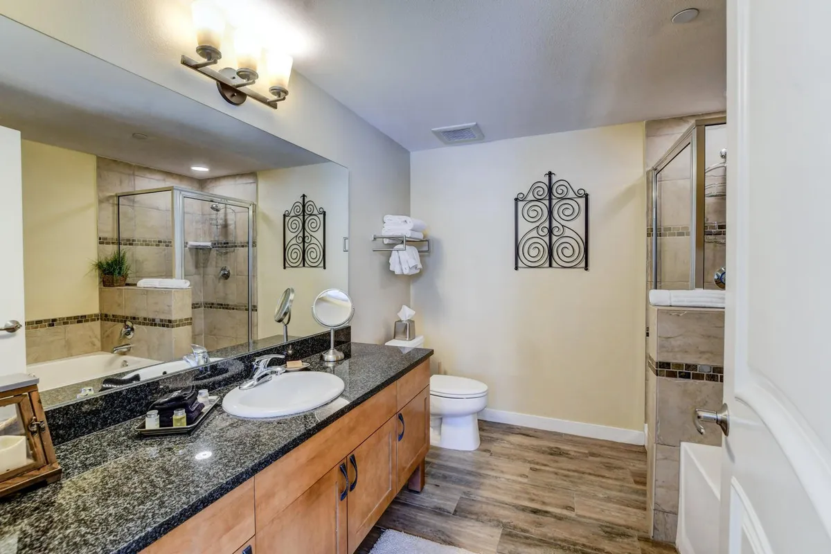 A modern bathroom featuring a granite countertop, illuminated by natural light, with a large mirror, a sleek shower enclosure, and decorative wall art. The space feels clean, spa-like, and inviting for self-care routines.