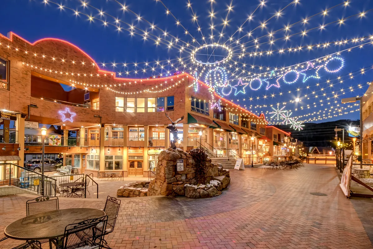An inviting village plaza comes to life with decorative string lights, outdoor seating, a stone fountain, and charming storefronts with awnings. The space is filled with energy, suggesting evening gatherings, dining, and socializing amidst a festive, welcoming atmosphere.