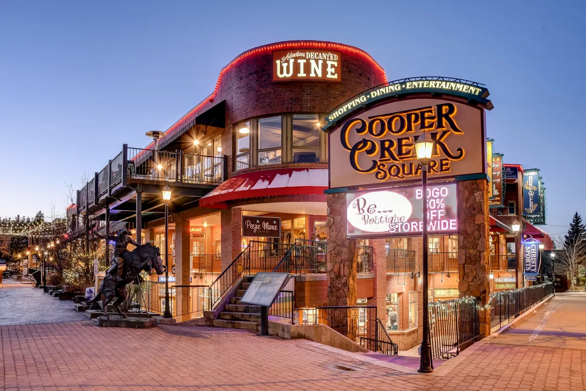 Cooper Creek Square is featured with its prominent sign, wine shop, outdoor dining area, and a whimsical statue. The scene is bustling yet intimate, with warm lighting suggesting social gatherings, culinary adventures, and shopping within steps of the accommodations.