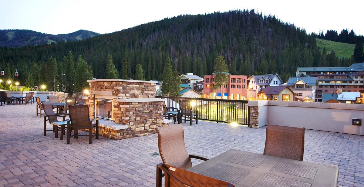 An outdoor seating area surrounds an elegant fireplace, with groups relaxing together as dusk settles over the mountains. The resort buildings provide a warm, inviting backdrop, and the scene is framed by lush trees and panoramic mountain views. The atmosphere is both cozy and sophisticated, ideal for unwinding after a day of adventure.