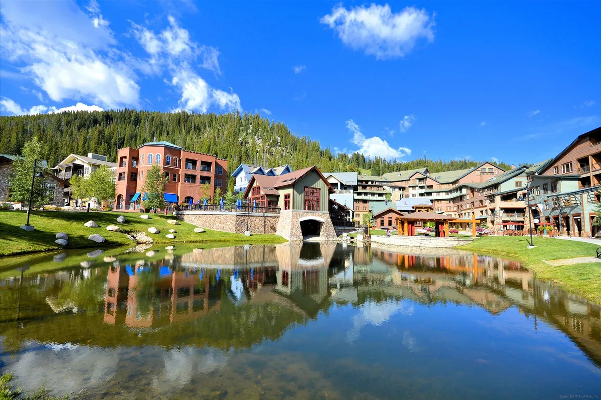 A cheerful exterior scene capturing colorful buildings reflected in a calm body of water, with green grass, trees, and a small bridge adding to the lively village atmosphere. Visitors stroll along the pathways, relax on the grass, and enjoy the vibrant resort village surroundings.