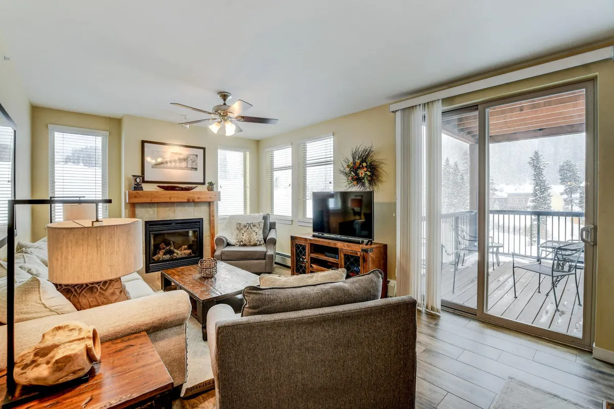 A close-up view of the living space features a plush sofa, a rustic stone fireplace, a coffee table topped with games, a smart TV mounted on the wall, and large windows revealing hints of the mountains outside. The scene is inviting, ideal for movie nights or relaxing after a day outdoors.