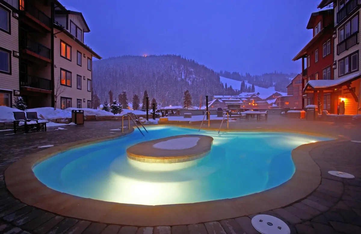The photo captures a vibrant outdoor plaza area featuring a large, illuminated hot tub surrounded by snow and cozy seating. Guests are visible enjoying the warm water and socializing, with the scenic, snow-covered landscape and glowing resort buildings in the background.