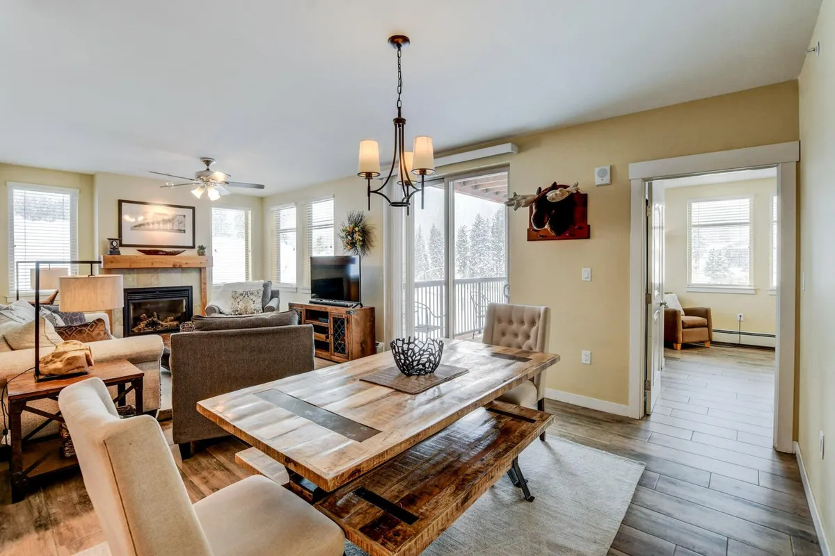 This inviting interior photograph captures a spacious open-concept living and dining area. The focal points include a central stone-clad gas fireplace, a wooden dining table set for gatherings, plush sofas oriented toward a wall-mounted smart TV, and expansive floor-to-ceiling windows that showcase sweeping mountain views. Natural light streams in, creating a warm, communal atmosphere perfect for both relaxation and entertainment.