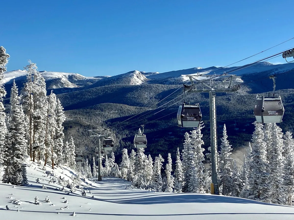 A scenic view captures a ski lift ascending over snow-covered slopes, surrounded by towering evergreen trees and distant mountain peaks. The scene suggests adventure and tranquility, with guests enjoying the outdoors and panoramic vistas.