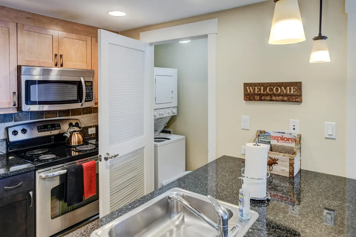 A close-up of the kitchen's granite countertop, modern appliances, and a visible welcome sign, with a cozy laundry area in the background. The image radiates warmth and functionality, highlighting thoughtful design details and the premium finishes.