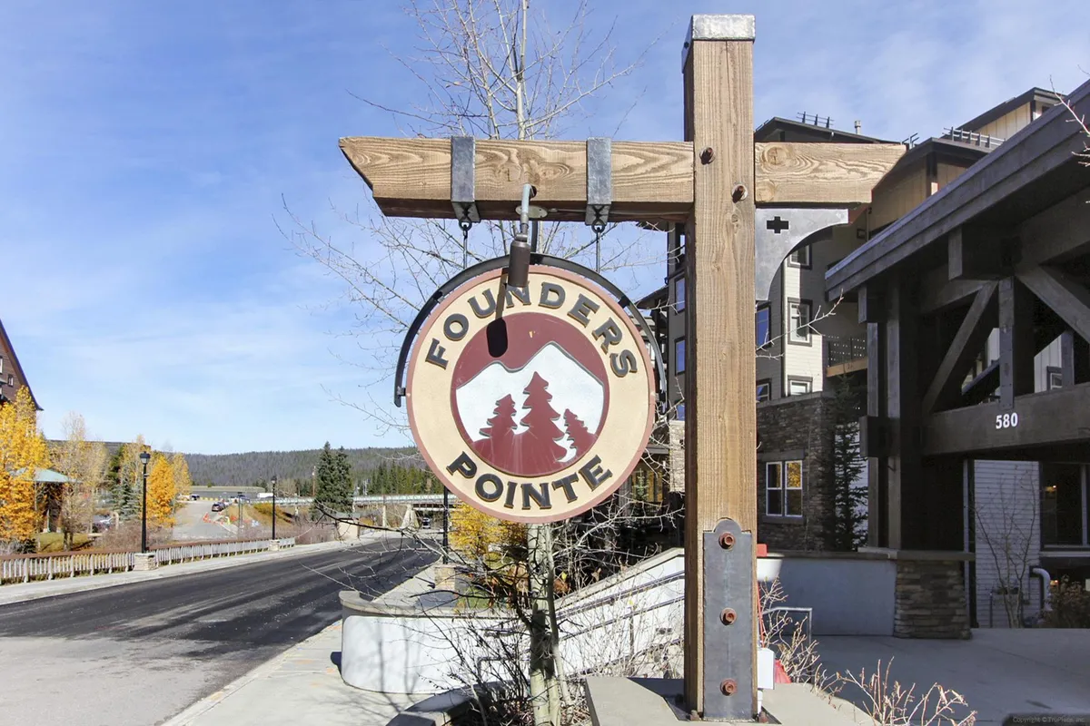 This exterior image showcases the Founder's Pointe sign set among surrounding trees and nearby rustic buildings. The scene is bathed in natural light, with architectural elements suggesting a welcoming, mountain-resort ambiance. The backdrop hints at the proximity to nature and the heart of the village, inviting guests to explore.
