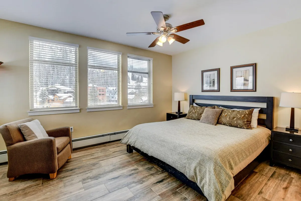 A tranquil bedroom with a comfortable bed, large windows letting in natural light, nightstands on either side, an armchair for reading, and decorative frames above the headboard. The bedding looks soft and inviting, contributing to a restful atmosphere.