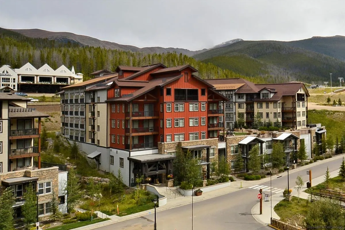 The condo's main building stands prominently against a backdrop of mountains and trees, with red architectural accents adding character. A sidewalk leads visitors through the landscaped grounds, hinting at a serene arrival experience.