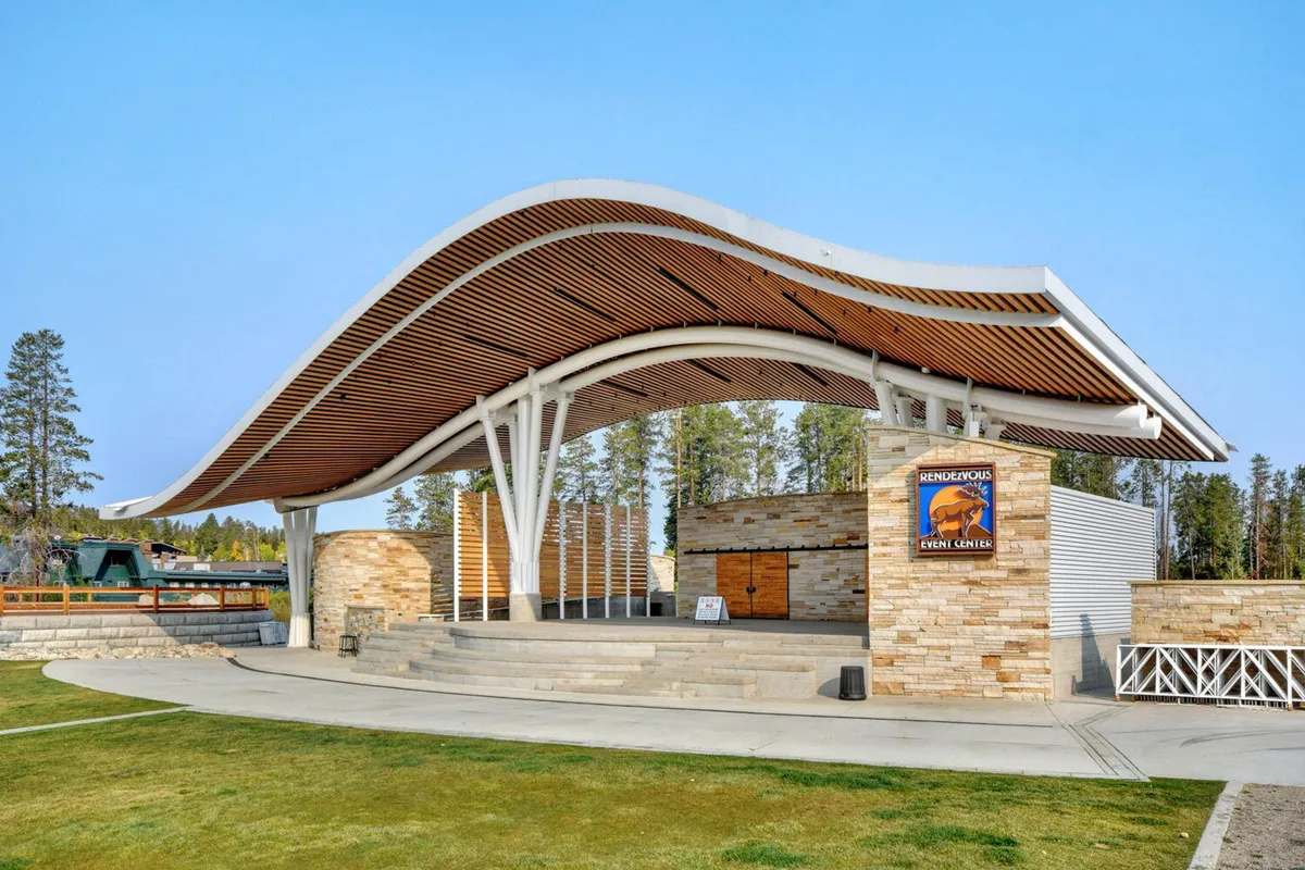 An exterior view of a modern, open-air venue with a distinctive curved roof, sturdy stone walls, and exposed wooden beams. The space is set up as an open stage area suitable for events and gatherings, with ample natural light and picturesque sunset views.